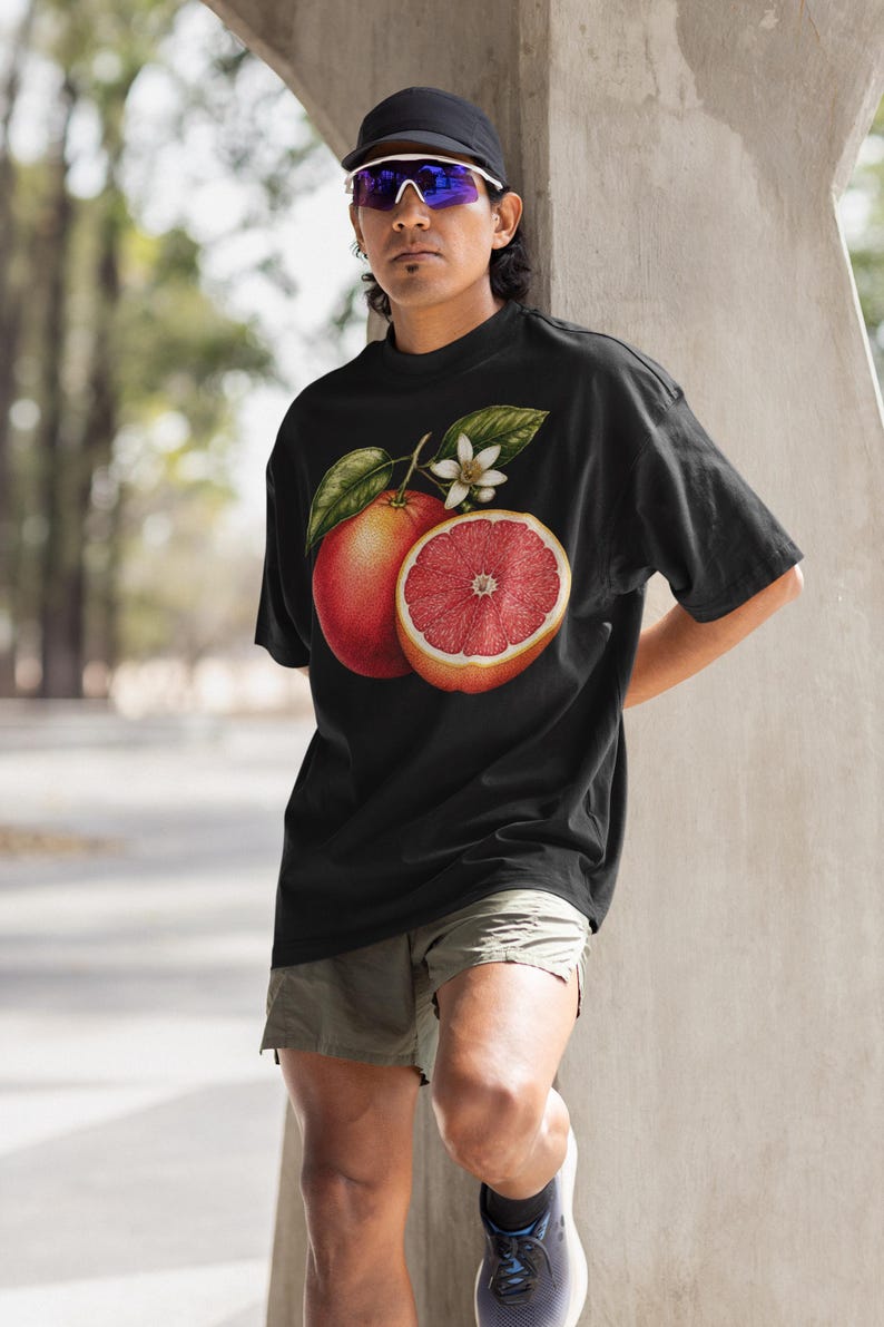 Puede incluir: Una camiseta negra de manga corta con un estampado gr&aacute;fico detallado de un pomelo entero y un pomelo cortado por la mitad con hojas verdes y una peque&ntilde;a flor blanca centrado en la zona del pecho de la prenda.