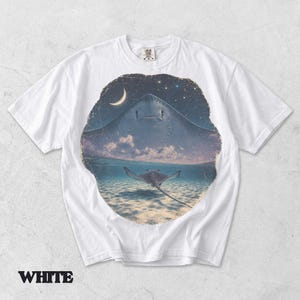 Pode incluir: Uma t-shirt branca de manga curta com uma impress&atilde;o gr&aacute;fica circular. O design mostra a silhueta de uma raia grande contra um c&eacute;u estrelado com uma lua crescente, transicionando para uma cena subaqu&aacute;tica com uma raia mais pequena.
