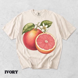 Puede incluir: Una camiseta de manga corta color marfil con una ilustraci&oacute;n bot&aacute;nica de un pomelo entero y medio pomelo con hojas verdes y una flor blanca. El gr&aacute;fico est&aacute; centrado en el pecho y la camiseta tiene un cuello redondo.