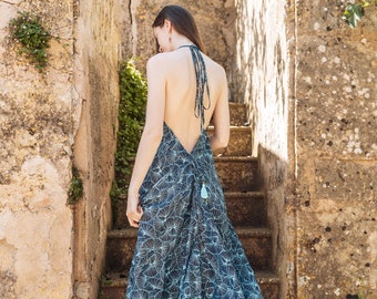 May include: A floor-length halter dress in a deep teal blue fabric featuring a repeating white fan-like geometric pattern. The dress has an open back design with thin tie straps at the neck and a small light blue tassel detail at the lower back.