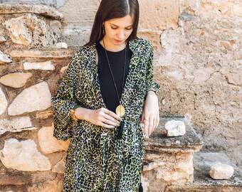 May include: A woman wearing a green and black leopard print kimono with a long black necklace and a gold pendant. She is standing in front of a stone wall.