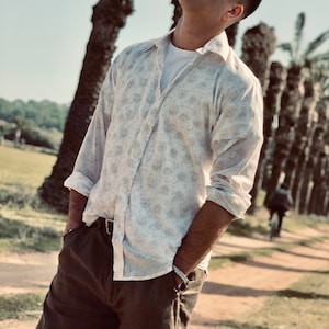 Op de afbeelding: Een man draagt een wit overhemd met knopen en een bloemenpatroon, een bruine broek en een zilveren armband. Hij staat in een palmenbos en kijkt omhoog.