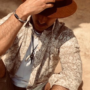 May include: A man wearing a brown fedora, a white shirt with a skull pattern, and a brown leather bracelet. He is also wearing a necklace with a bone pendant.