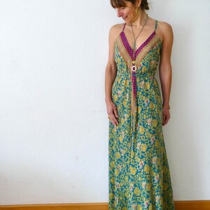 May include: A woman wearing a long, blue and yellow floral print maxi dress with a pink and gold trim. The dress has a V-neckline and thin straps. She is wearing a long gold necklace with a turquoise stone pendant.