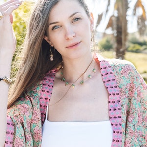 May include: A woman wearing a floral kimono with pink and teal patterns, a white top, and gold jewelry. The jewelry includes a necklace with green and gold accents and earrings with pink stones. The background features palm trees and sunlight.