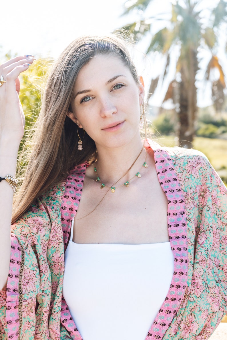 May include: A woman wearing a floral kimono with pink and teal patterns, a white top, and gold jewelry. The jewelry includes a necklace with green and gold accents and earrings with pink stones. The background features palm trees and sunlight.