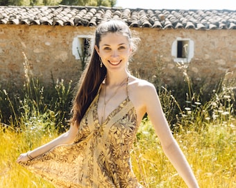 May include: A woman wearing a yellow and brown floral dress smiles while standing in a field of long grass.