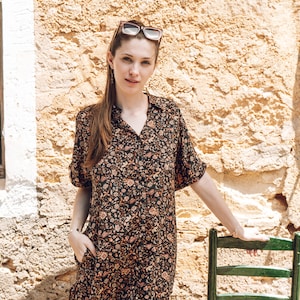 Può includere: Una donna con un vestito a stampa floreale e occhiali da sole si trova in piedi davanti a un muro di pietra. Si appoggia a una sedia di legno verde con un sedile di paglia.