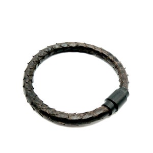 Puede incluir: Un brazalete de cuero marrón oscuro con una superficie texturizada y un cierre de plástico negro.