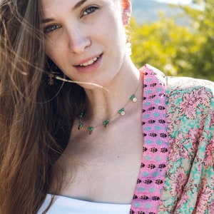May include: A woman wearing a gold necklace with green and gold charms, and matching earrings. She is also wearing a white top and a pink and turquoise patterned kimono. The background is blurred with green and yellow tones.