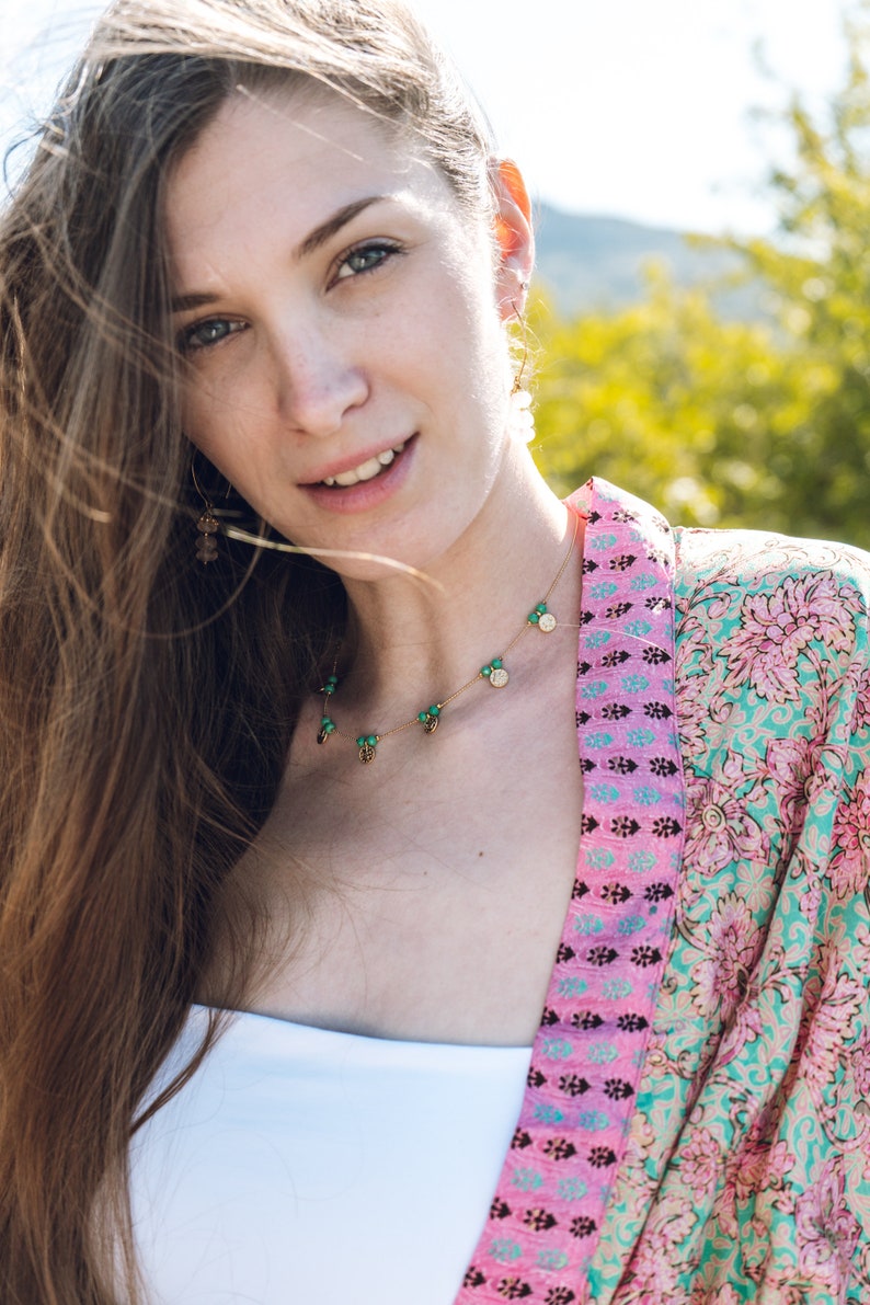 May include: A woman wearing a gold necklace with green and gold charms, and matching earrings. She is also wearing a white top and a pink and turquoise patterned kimono. The background is blurred with green and yellow tones.