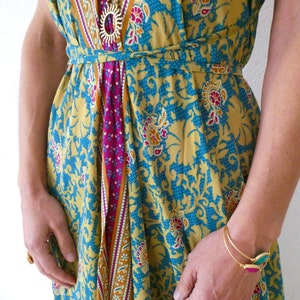 May include: A yellow, blue, and pink patterned maxi dress with a gold chain necklace and a gold bracelet. The dress has a tie at the waist.