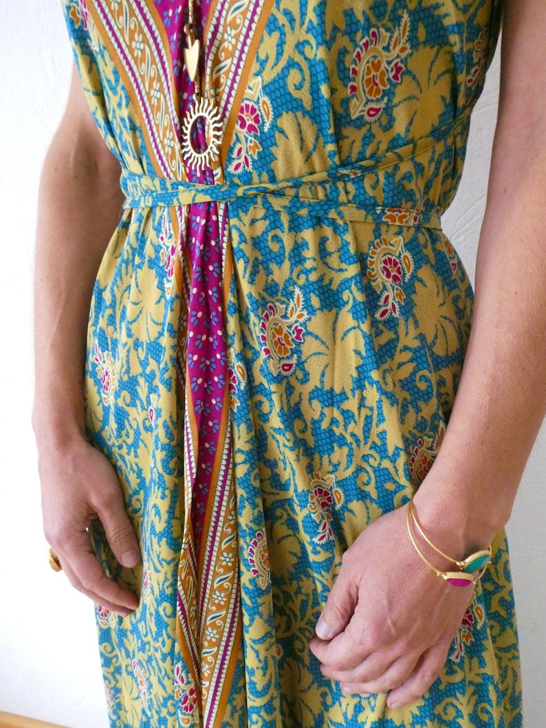 May include: A yellow, blue, and pink patterned maxi dress with a gold chain necklace and a gold bracelet. The dress has a tie at the waist.