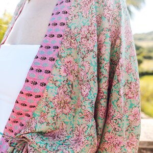 May include: A floral print kimono in shades of teal, pink, and gold. The kimono features a large floral pattern and a contrasting pink border. The garment is tied at the waist and appears to be made of a lightweight fabric.