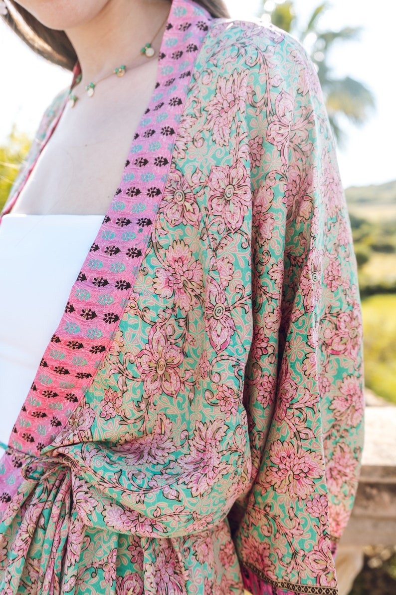 May include: A floral print kimono in shades of teal, pink, and gold. The kimono features a large floral pattern and a contrasting pink border. The garment is tied at the waist and appears to be made of a lightweight fabric.