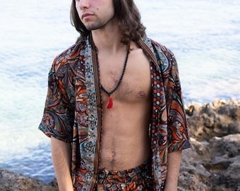 Puede incluir: Un hombre con una camisa de manga corta con estampado marrón y azul y pantalones cortos a juego. Está de pie en una playa rocosa con el mar de fondo. Lleva un collar de cuentas negras con un borla roja.