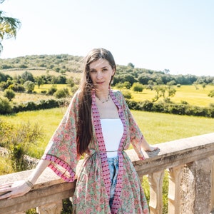 May include: A floral print kimono in shades of pink, green, and purple. The lightweight garment is worn over a white top and blue jeans. The kimono features a tie closure and wide sleeves. The background shows a sunny outdoor scene.