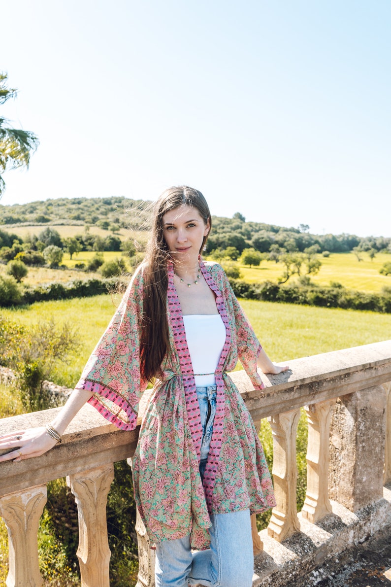 May include: A floral print kimono in shades of pink, green, and purple. The lightweight garment is worn over a white top and blue jeans. The kimono features a tie closure and wide sleeves. The background shows a sunny outdoor scene.