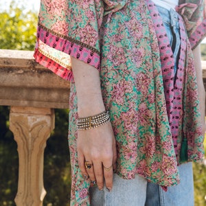 May include: A floral print kimono in shades of teal, pink, and gold. The kimono is paired with light wash denim jeans and a white top. A multi-strand bracelet and gold rings adorn the wrist. The kimono's design features a repeating floral pattern.