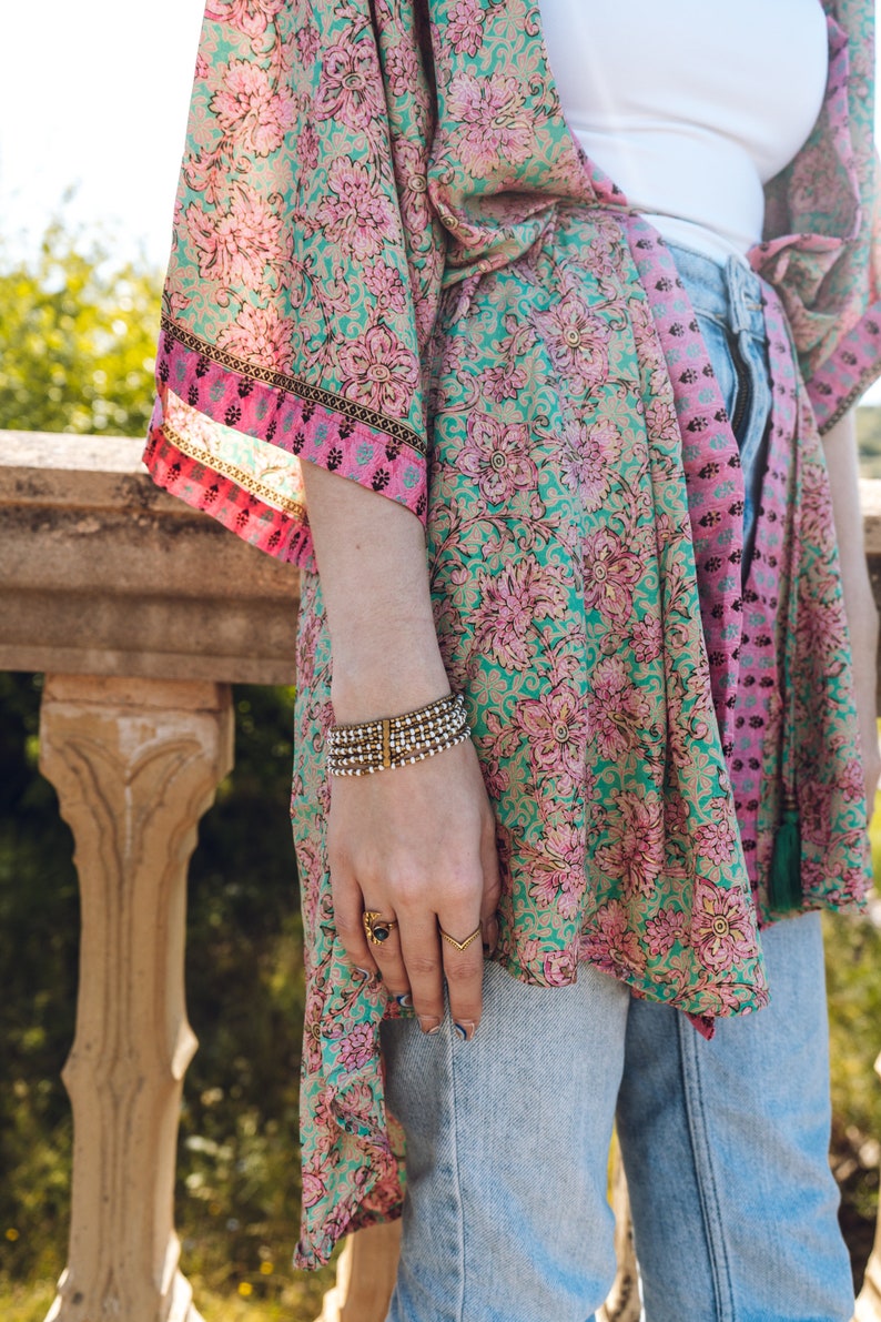 May include: A floral print kimono in shades of teal, pink, and gold. The kimono is paired with light wash denim jeans and a white top. A multi-strand bracelet and gold rings adorn the wrist. The kimono's design features a repeating floral pattern.