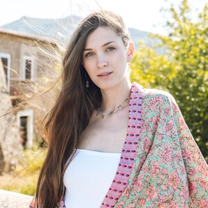 May include: A woman wearing a floral print kimono in shades of pink and teal, paired with a white top and denim jeans. She is also wearing a necklace and earrings. The background features a stone building and greenery.