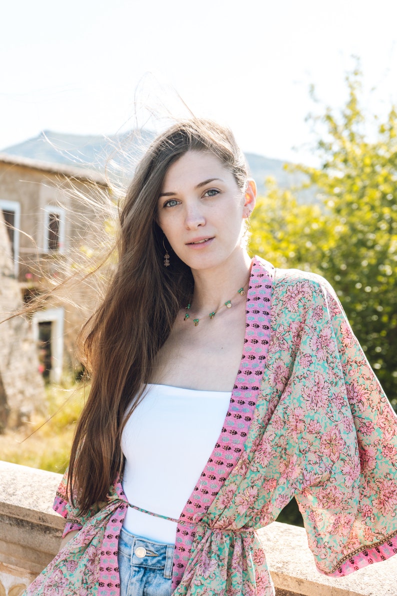 May include: A woman wearing a floral print kimono in shades of pink and teal, paired with a white top and denim jeans. She is also wearing a necklace and earrings. The background features a stone building and greenery.
