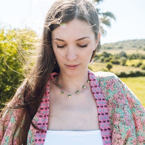 May include: A woman wearing a pink floral kimono and a delicate necklace with green and gold charms. The necklace is the focal point, complemented by the kimono's vibrant colors and the natural outdoor setting.