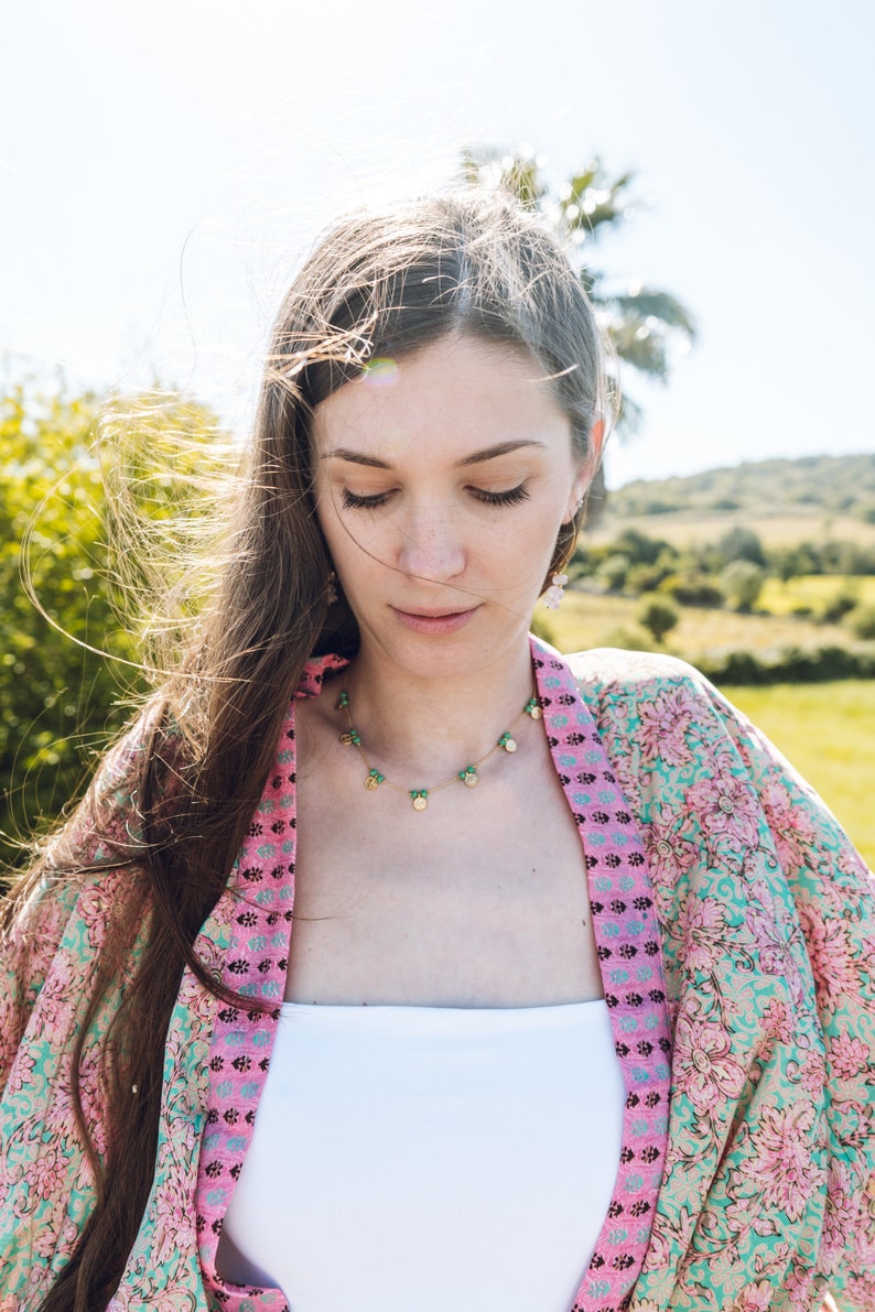 May include: A woman wearing a pink floral kimono and a delicate necklace with green and gold charms. The necklace is the focal point, complemented by the kimono's vibrant colors and the natural outdoor setting.