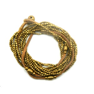 Puede incluir: Un brazalete dorado de envoltura con múltiples hebras de pequeñas cuentas cuadradas y un cordón de cuero marrón.