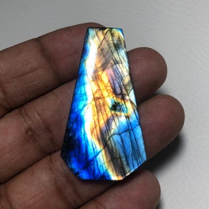 Könnte beinhalten: Ein blauer und goldener Labradorit-Cabochon mit irisierendem Schimmer. Der Edelstein ist in Tropfenform geschliffen und hat eine glatte, polierte Oberfläche.