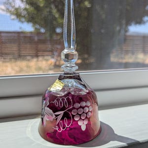 Può includere: Una campana in vetro trasparente e magenta intenso con un manico alto e sottile dalla base sfaccettata. Il corpo della campana è decorato con grappoli d'uva e motivi a vite incisi in bianco, e include un piccolo adesivo color oro.