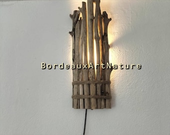 May include: A wall-mounted light fixture constructed from vertical driftwood branches bound together with natural twine. The lamp emits a warm, glowing light from behind the wood. The text BordeauxArtNature is centered over the fixture.