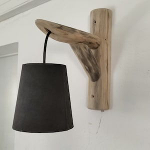 Peut inclure: Un luminaire mural en bois naturel avec un abat-jour en tissu noir. Le luminaire est monté sur un mur blanc.