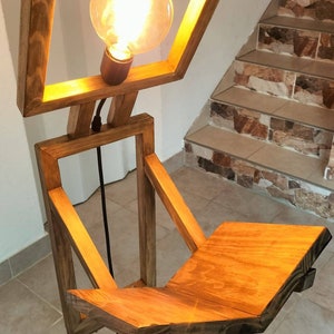 Peut inclure: Un lampadaire en bois avec un luminaire carr&eacute; et une &eacute;tag&egrave;re. La lampe est en bois naturel et a un aspect chaleureux et rustique. Le luminaire est &eacute;clair&eacute; par une ampoule, projetant une lueur chaude.