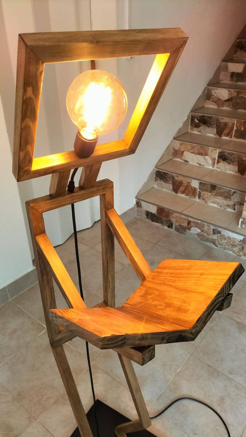 Peut inclure: Un lampadaire en bois avec un luminaire carr&eacute; et une &eacute;tag&egrave;re. La lampe est en bois naturel et a un aspect chaleureux et rustique. Le luminaire est &eacute;clair&eacute; par une ampoule, projetant une lueur chaude.
