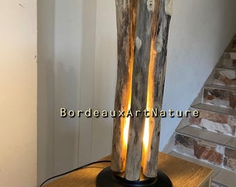 May include: A floor lamp featuring three vertical pieces of weathered driftwood arranged around a central light source, mounted on a circular black metal base. The text BordeauxArtNature is visible across the center of the lamp structure.