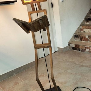 Peut inclure: Un lampadaire en bois en forme de personne tenant un livre. La lampe a un luminaire carr&eacute; avec une ampoule &agrave; l'int&eacute;rieur. La lampe est sur une base noire et a un cordon noir avec un interrupteur au pied.