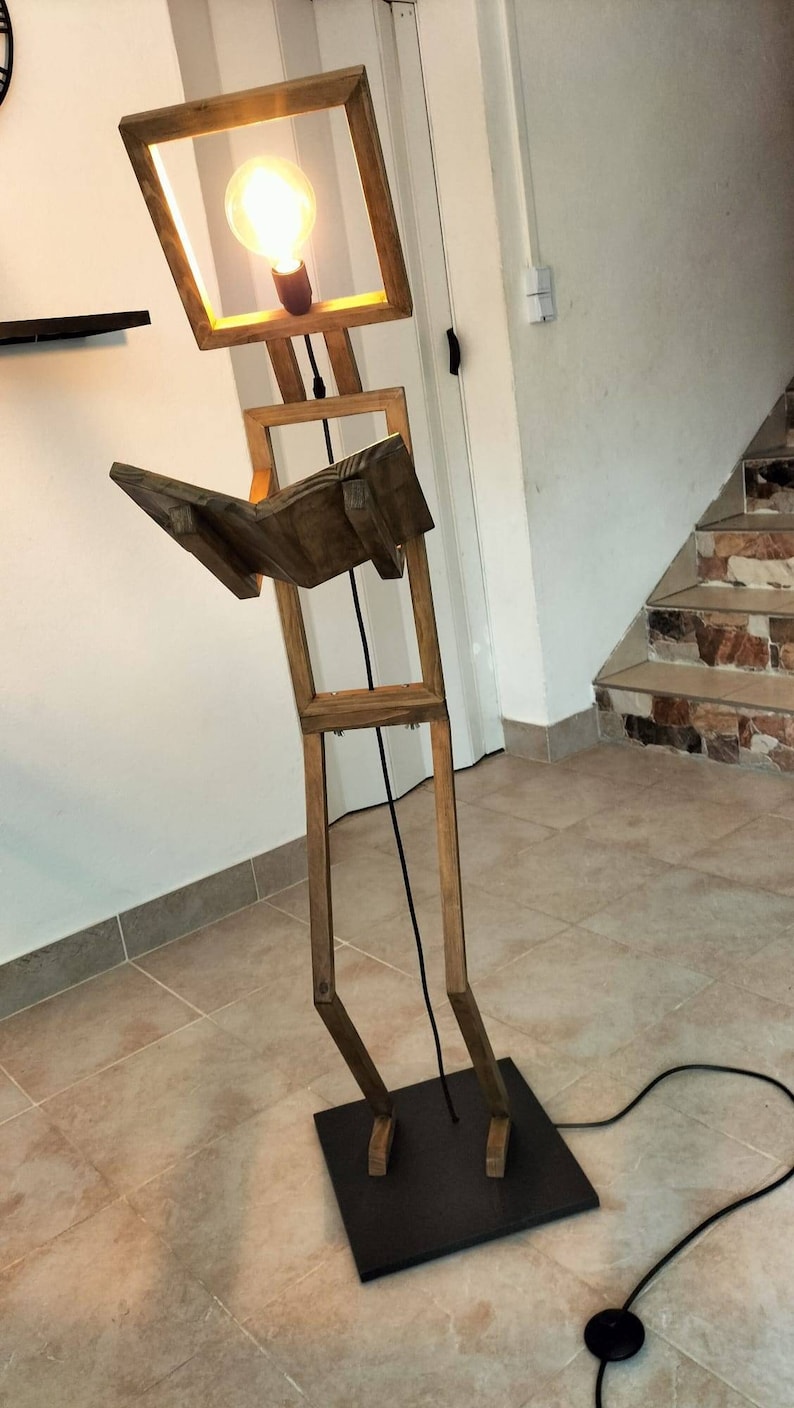 Peut inclure: Un lampadaire en bois en forme de personne tenant un livre. La lampe a un luminaire carr&eacute; avec une ampoule &agrave; l'int&eacute;rieur. La lampe est sur une base noire et a un cordon noir avec un interrupteur au pied.