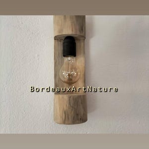 May include: A wall-mounted light fixture featuring a cylindrical piece of light-colored natural wood with a vertical cutout. A black socket holds a clear glass light bulb inside the cutout. The text BordeauxArtNature is printed on the wood.