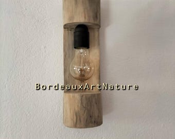 May include: A wall-mounted light fixture featuring a cylindrical piece of light-colored natural wood with a vertical cutout. A black socket holds a clear glass light bulb inside the cutout. The text BordeauxArtNature is printed on the wood.