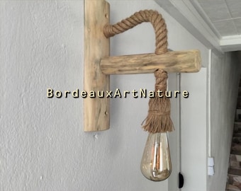 May include: A wall-mounted light fixture featuring a vertical light-colored wooden plank, a horizontal wooden beam, and a thick tan rope. The rope wraps around the beam and ends in a frayed tassel holding a teardrop-shaped light bulb. Text reads BordeauxArtNature.