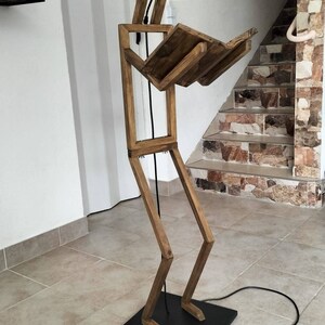 Peut inclure: Un lampadaire en bois en forme de personne lisant un livre. La lampe a une ampoule dans la t&ecirc;te et un cordon noir.