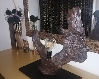 May include: A unique table lamp made from a piece of driftwood with a black base. The lamp has a single light bulb and is lit on.