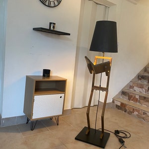 May include: A wooden floor lamp shaped like a person reading a book. The lamp has a black lampshade and a black base. The lamp is next to a small wooden cabinet with a white door.