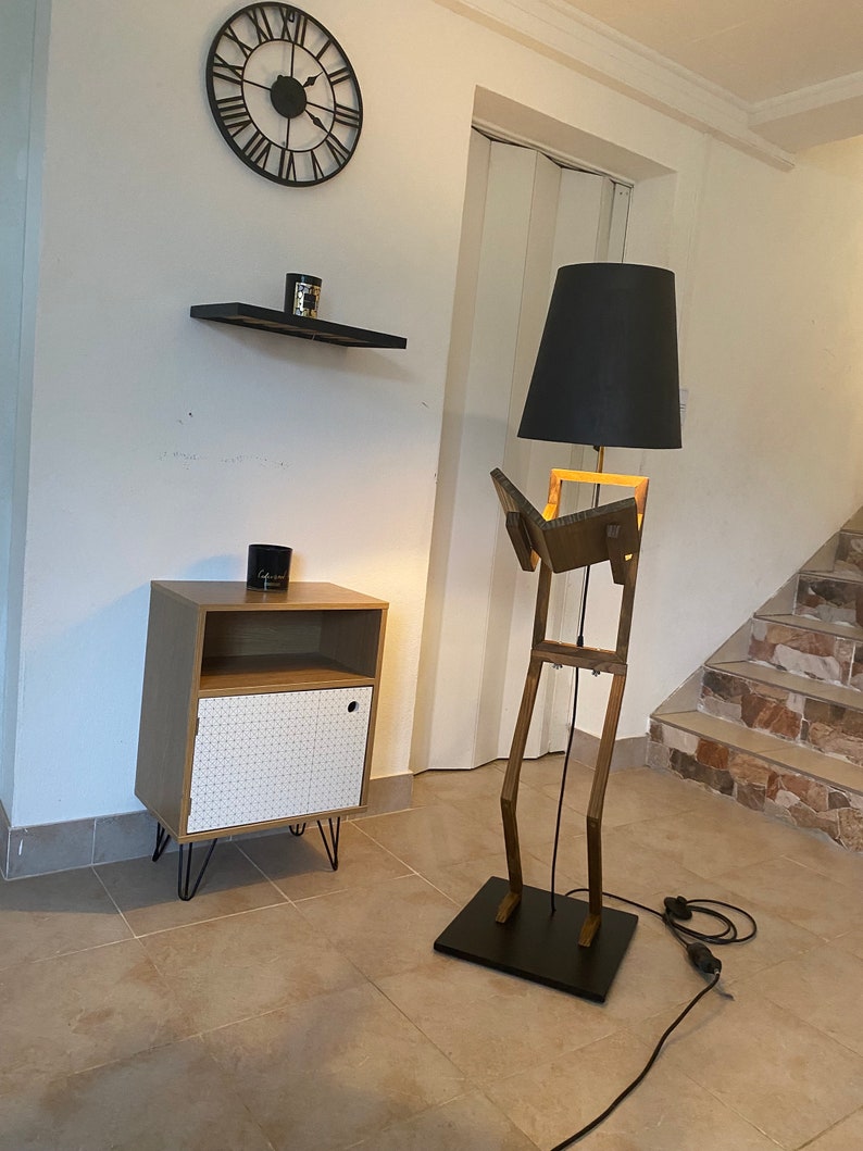 May include: A wooden floor lamp shaped like a person reading a book. The lamp has a black lampshade and a black base. The lamp is next to a small wooden cabinet with a white door.