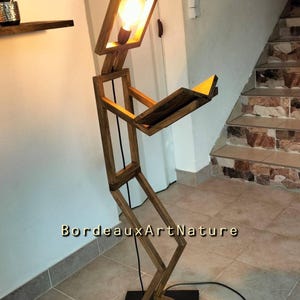 May include: A floor lamp constructed from dark stained wood, shaped like a human figure standing and holding an open book. The head of the figure is a square frame containing a glowing light bulb. The text BordeauxArtNature is visible.