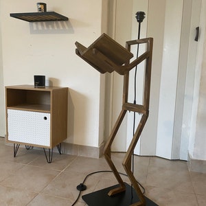 May include: A wooden floor lamp shaped like a person with a shelf for a book. The lamp has a black cord and a black base.