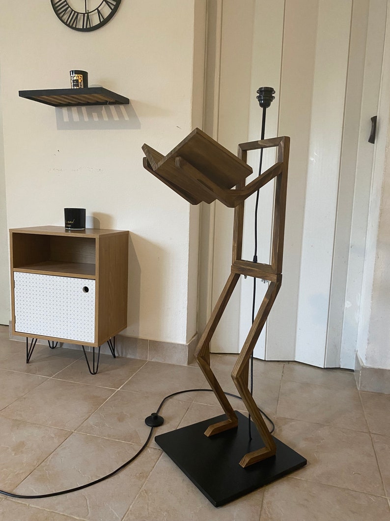 May include: A wooden floor lamp shaped like a person with a shelf for a book. The lamp has a black cord and a black base.