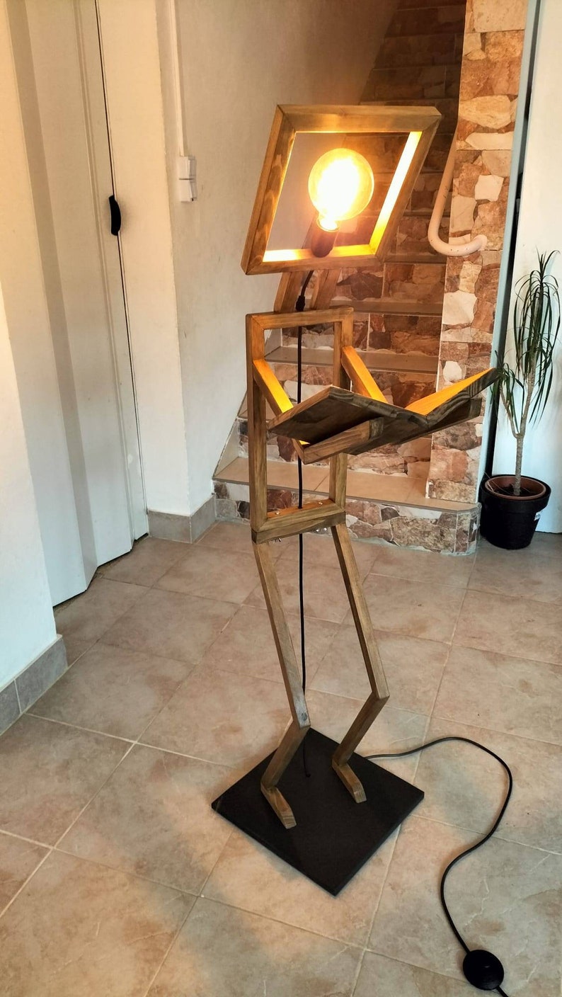 Peut inclure: Un lampadaire en bois en forme de personne lisant un livre. La lampe a une ampoule dans la t&ecirc;te et un cordon noir avec un interrupteur au pied.