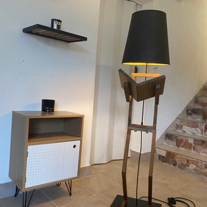 May include: A wooden floor lamp with a black lampshade. The lamp is shaped like a person with a long neck and legs. The lamp is standing on a black base.